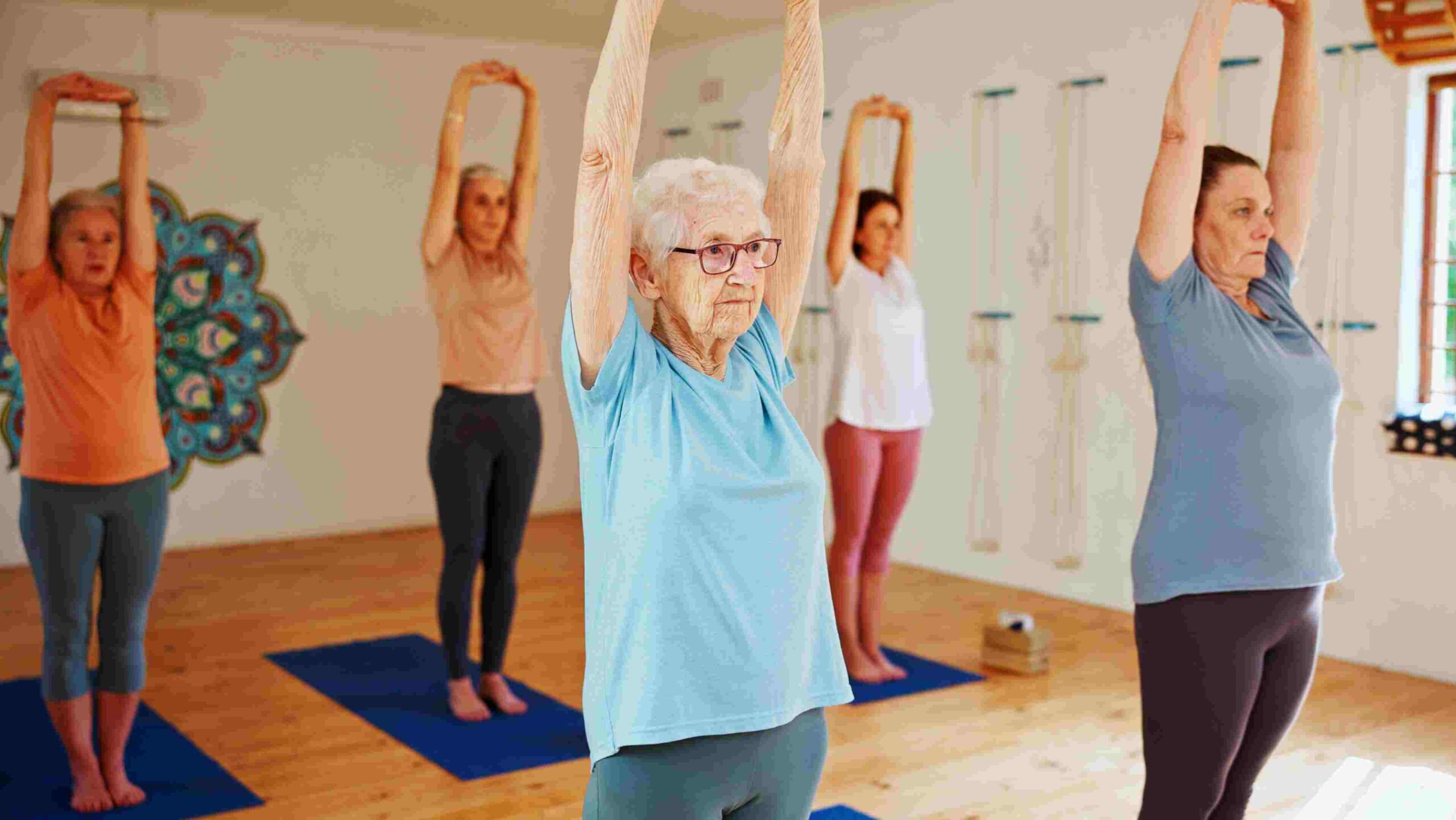 Group of senior women in a gym standing and stretching on yoga mats.