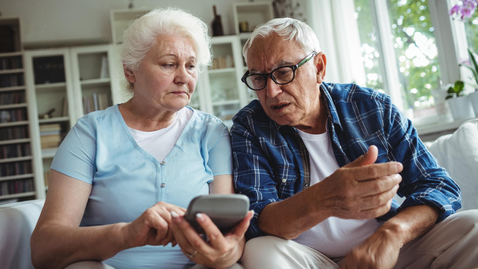 Two seniors looking worried while sitting on a couch using a calculator.